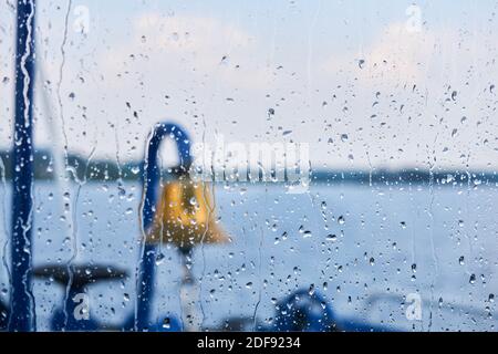 Hintergrund - Regentropfen auf dem Glas des Steuerhauses, hinter dem man eine verschwommene Glocke, andere Details des Schiffes und eine regnerische Wasserlandschaft erahnen kann Stockfoto