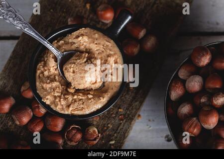 Haselnussbutter in Keramikbecher mit geschälten Haselnüssen auf Holzgrund. Stockfoto