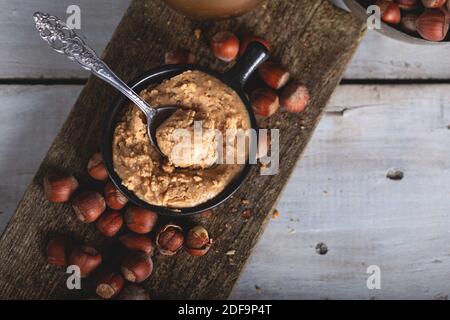 Haselnussbutter in Keramikbecher mit geschälten Haselnüssen auf Holzgrund. Stockfoto