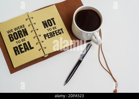 Inspirierende und Motivation Leben Zitat. Notizblock und Kaffeetasse auf weißem Hintergrund - Wir wurden geboren, um echt zu sein, nicht um perfekt zu sein. Stockfoto