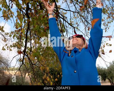 Ein flacher Fokus einer fröhlichen mittleren Alters blonde Frau werfen Die gefallenen Blätter in der Luft Stockfoto