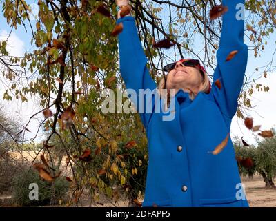 Ein flacher Fokus einer fröhlichen mittleren Alters blonde Frau werfen Die gefallenen Blätter in der Luft Stockfoto