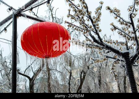 Eine chinesische rote Laterne hängt im Winter auf der Straße, es ist sehr eisig, Eiszapfen hängen. Vor dem Hintergrund von Glühbirnen in der Stadt. Stockfoto