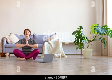 Eine ältere Frau praktiziert Yoga. Training zu Hause, geleitet von einem Online-Trainer. Lotus-Pose. Stockfoto