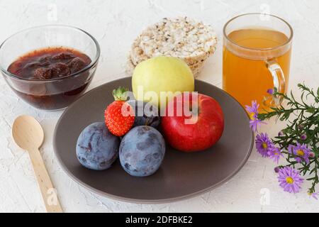 Gepflückte reife Pflaumen, Äpfel, eine Erdbeere auf dem Teller und eine Glasschüssel mit Marmelade, gepuffte Reiskuchen, eine Tasse Tee auf dem weiß strukturierten Hintergrund. Ju Stockfoto