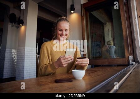 Junge Frau, die Online-Zahlung mit Karte und Smartphone, während Sitzen und Kaffee im Café genießen Stockfoto