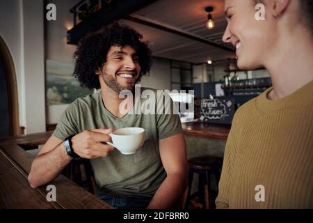 Schöner junger Mann mit lockigen Haaren hält Kaffeetasse sitzen Mit schöner Frau und im Café reden Stockfoto