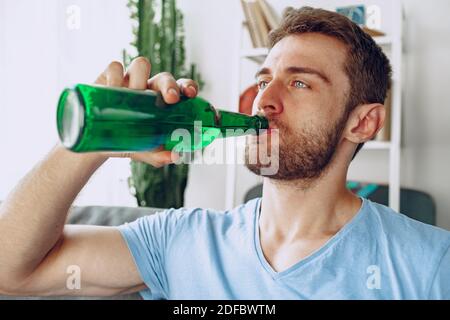 Bärtiger Mann, der Bier aus der Flasche trinkt, während er darauf sitzt Sofa zu Hause Stockfoto