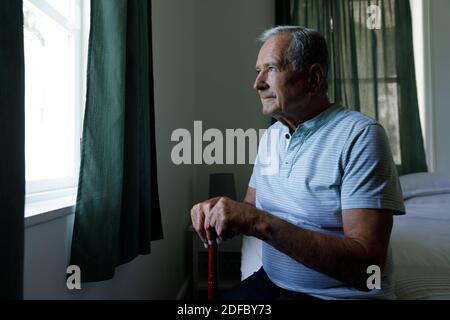 Älterer kaukasischer Mann, der einen Spazierstock hält und aus dem Fenster schaut Zu Hause Stockfoto