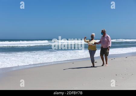 Ältere kaukasische Paar mit Gesichtsmasken auf Strand zu Fuß reden Stockfoto