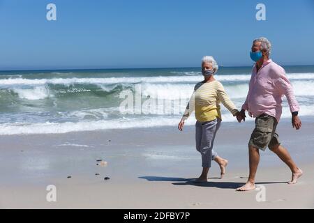 Ältere kaukasische Pärchen mit Gesichtsmasken am Strand halten Hände Stockfoto