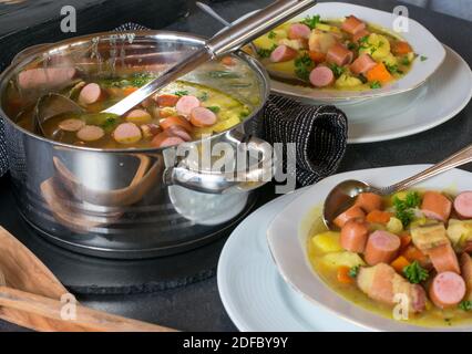 Deutsche Kartoffelsuppe mit Hot Dog und Gemüse auf einem Tisch mit Topf, Schöpfkelle und Teller serviert Stockfoto