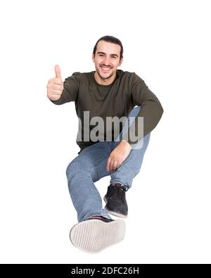 Zufriedener, zwangloser junger Mann, der auf dem Boden sitzt, lächelt zur Kamera und zeigt den Daumen nach oben. Lifestyle voller Länge Porträt glücklich Student Kerl Stockfoto