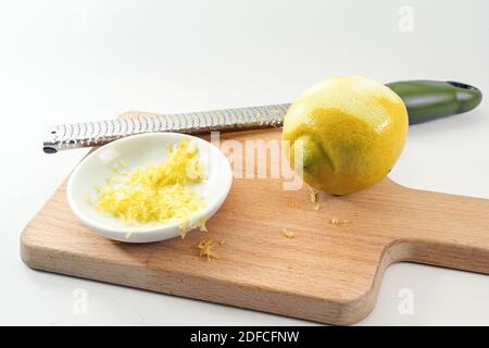 Geriebene Zitronenschale in einer kleinen Schüssel, Obst und Reibe auf einem hölzernen Küchentafel, heller Hintergrund, Kopierraum, ausgewählter Fokus, enge Schärfentiefe Stockfoto
