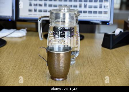 Eine Nahaufnahme einer Kupfertasse mit einem Glaskrug Gefüllt mit Wasser auf einer hölzernen Oberfläche Stockfoto