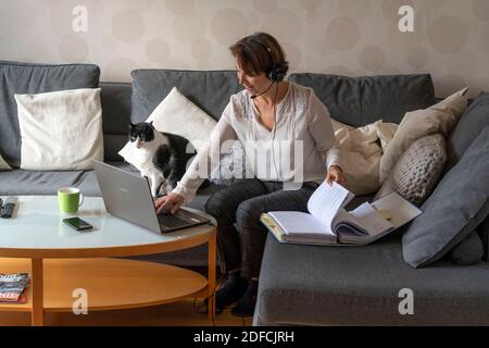 Frau, Mitte 50, arbeitet von zu Hause, mit Laptop und kommuniziert über Headset mit Kollegen, Home Office, auf dem Sofa, Stockfoto