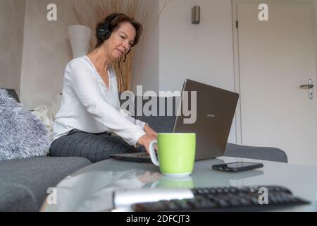 Frau, Mitte 50, arbeitet von zu Hause, mit Laptop und kommuniziert über Headset mit Kollegen, Home Office, auf dem Sofa, Stockfoto