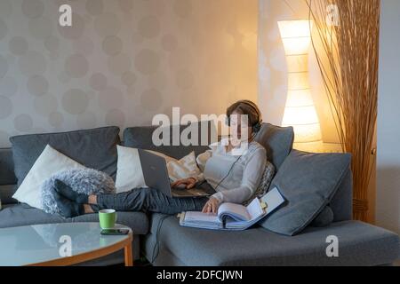 Frau, Mitte 50, arbeitet von zu Hause, mit Laptop und kommuniziert über Headset mit Kollegen, Home Office, auf dem Sofa, Stockfoto