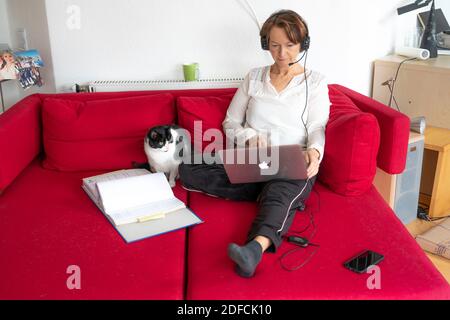 Frau, Mitte 50, arbeitet von zu Hause, mit Laptop und kommuniziert über Headset mit Kollegen, Home Office, auf dem Sofa, Stockfoto