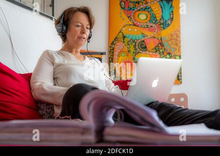Frau, Mitte 50, arbeitet von zu Hause, mit Laptop und kommuniziert über Headset mit Kollegen, Home Office, auf dem Sofa, Stockfoto