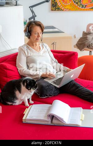 Frau, Mitte 50, arbeitet von zu Hause, mit Laptop und kommuniziert über Headset mit Kollegen, Home Office, auf dem Sofa, Stockfoto