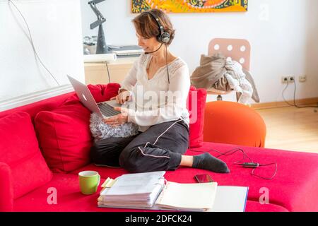 Frau, Mitte 50, arbeitet von zu Hause, mit Laptop und kommuniziert über Headset mit Kollegen, Home Office, auf dem Sofa, Stockfoto