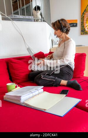 Frau, Mitte 50, arbeitet von zu Hause, mit Laptop und kommuniziert über Headset mit Kollegen, Home Office, auf dem Sofa, Stockfoto