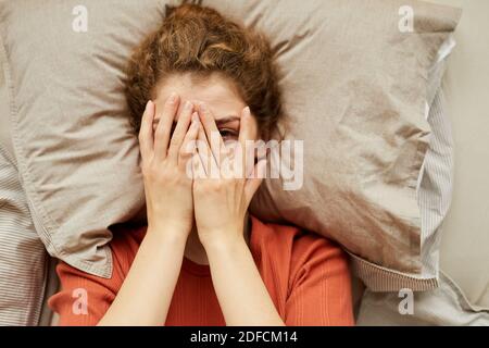 High-Angle-Ansicht der jungen Frau Blick auf die Kamera während Bedeckt ihr Gesicht mit den Händen, während sie im Bett ruht Stockfoto