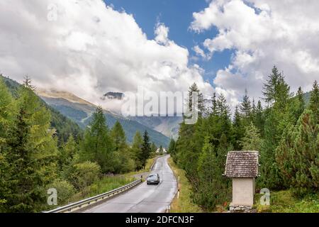 Ein Auto fährt entlang einer Bergstraße mit einem Votivschrein an der Seite unter einem dramatischen Himmel, Solda, Südtirol, Italien Stockfoto