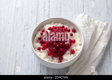 Reispudding mit Preiselbeermarmelade und frischen Preiselbeeren in einer weißen Schüssel auf hellblauem Hintergrund, selektiver Fokus. Köstliches traditionelles Frühstück Stockfoto