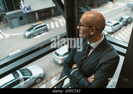 Ansicht von oben. Kontemplativer kahler Geschäftsmann in Anzug und Brille, die weg in das Fenster oder Glasaufzug mit einem geschäftigen Blick auf die Straße schaut. Nachdenklich Stockfoto