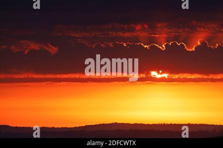 Strahlen von warmem orangefarbenem Licht brachen hinter einem Band aus flauschiger Wolke aus, während die Sonne zu einem in goldgelber Farbe getauchten Horizont untergeht. Stockfoto