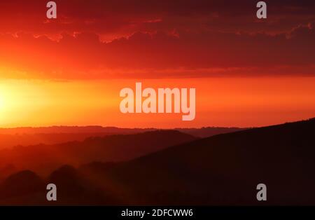 Die untergehende Sonne füllt den Himmel mit warmen orangefarbenen Farben, während Sonnenstrahlen über die schattigen, rollenden Schichten der Dorset-Landschaft fallen. Stockfoto