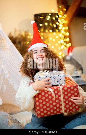 Hübsche Dame Frau hält große Weihnachten geschenkboxen in Armen tragen Strickpullover und weihnachtsmütze auf Bokeh-Lichtern Hintergrund Stockfoto