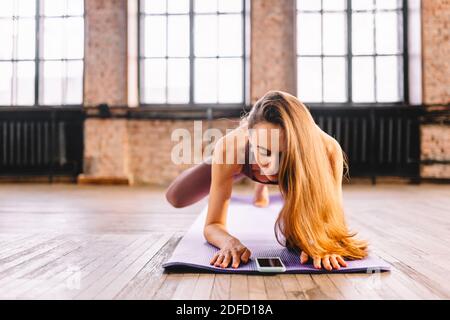 Junge langhaarige Frau tun Komplex von Stretching Yoga Asanas im Loft-Stil sonnigen Klasse mit ihrem Smartphone. Online-Unterricht und Training Stockfoto