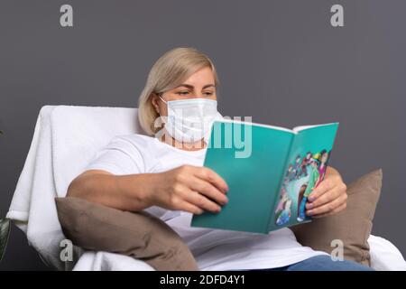 Eine Frau in einer medizinischen Maske liest Lieblingsbuch zu Hause in bequemen Stuhl. Stockfoto