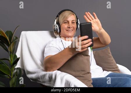 Ältere Frau mit Kopfhörern, Videoanruf am Telefon, zu Hause im Stuhl sitzend. Stockfoto