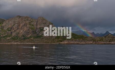 Kleines Boot vor der felsigen Küste der Austvagoya Insel, Lofoten, Norwegen, Skandinavien mit schroffen Bergen und spektakulären Regenbogen am Sommertag. Stockfoto