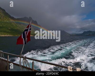 Norwegische Nationalflagge, die im Wind auf dem Heck des Touristenbootes mit majestätischem farbenfrohen Regenbogen über Bergen an der felsigen Küste Norwegens fliegt. Stockfoto