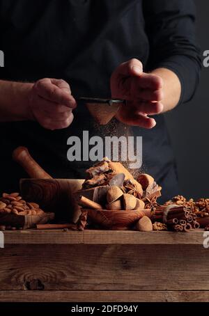 Der Koch streut die Schokolade mit Kakaopulver. Auf einem alten Holztisch Süßigkeiten, Kaffeebohnen, Zimt, Anis, Nüsse, und Stücke von gebrochenem schwarzem Choco Stockfoto