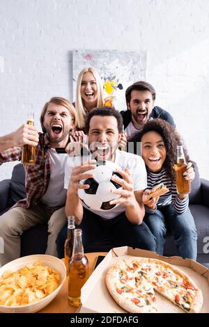 Aufgeregte multiethnische Fußballfans schreien, während sie Meisterschaft in der Nähe von Bier, Pizza und Chips beobachten Stockfoto