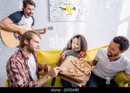 Multikulturelle Freunde essen Pizza auf dem Sofa, während junger Mann spielt Akustische Gitarre Stockfoto