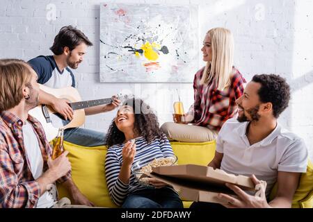 Junger Mann spielt akustische Gitarre zu fröhlichen multikulturellen Freunden während Party Stockfoto