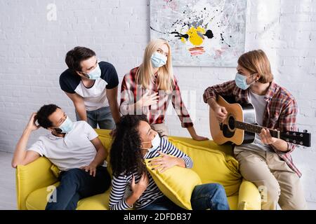 Junger Mann spielt akustische Gitarre zu multikulturellen Freunden in der Medizin Masken sitzen auf dem Sofa während der Party Stockfoto