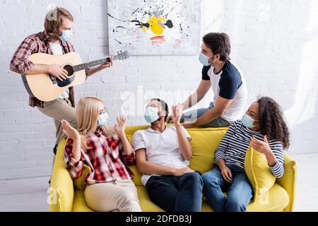 Junger Mann spielt akustische Gitarre, um multikulturelle Freunde sitzen auf Sofa in medizinischen Masken Stockfoto