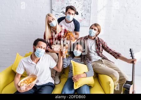 Multiethnische Freunde in medizinischen Masken Blick auf die Kamera beim Klirren Flaschen Bier während der Party Stockfoto