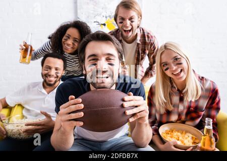 Aufgeregt Mann hält Rugby Ball während der Meisterschaft in der Nähe multiethnisch Freunde mit gemalten Gesichtern auf unscharfem Hintergrund Stockfoto