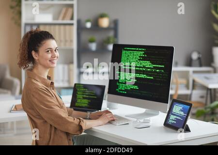 Porträt einer jungen Frau, die beim Sitzen auf die Kamera lächelt Der Tisch vor dem Computer-Monitor und arbeiten Mit Software Stockfoto
