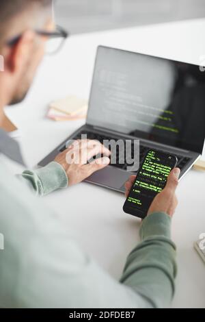 Rückansicht des Mannes, der am Tisch sitzt und programmiert Software auf Laptop und Handy Stockfoto