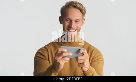 Attraktive aufgeregt bärtigen Mann schauen glücklich spielen im Spiel auf Smartphone und lächeln auf weißem Hintergrund Stockfoto